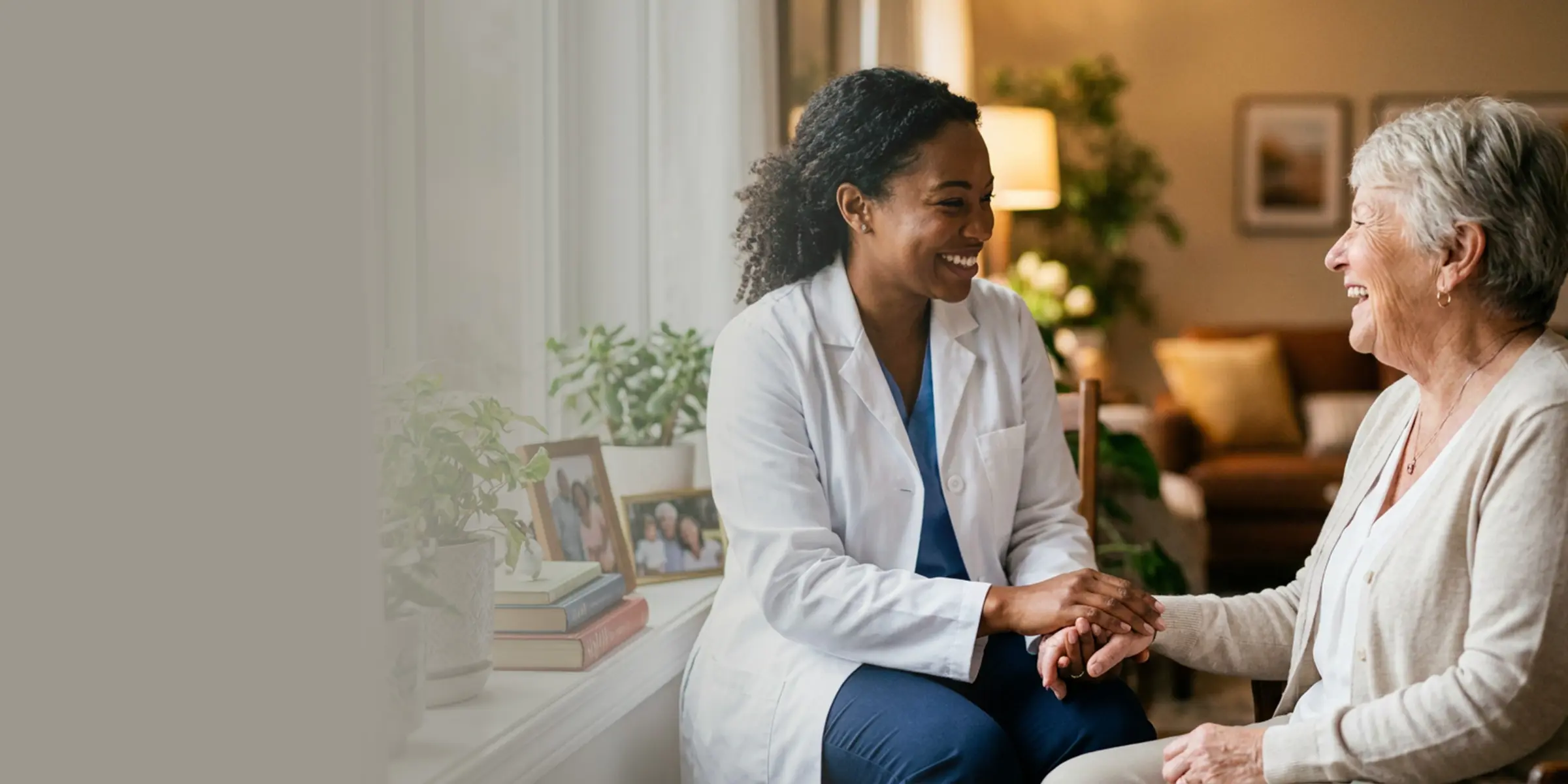 A clinician in a white coat holds hands and laughs with an older woman in her living room